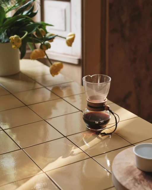 Pour-over coffee maker with flowers on a tiled table in a warm, inviting setting, showcasing a blend of functionality and style.