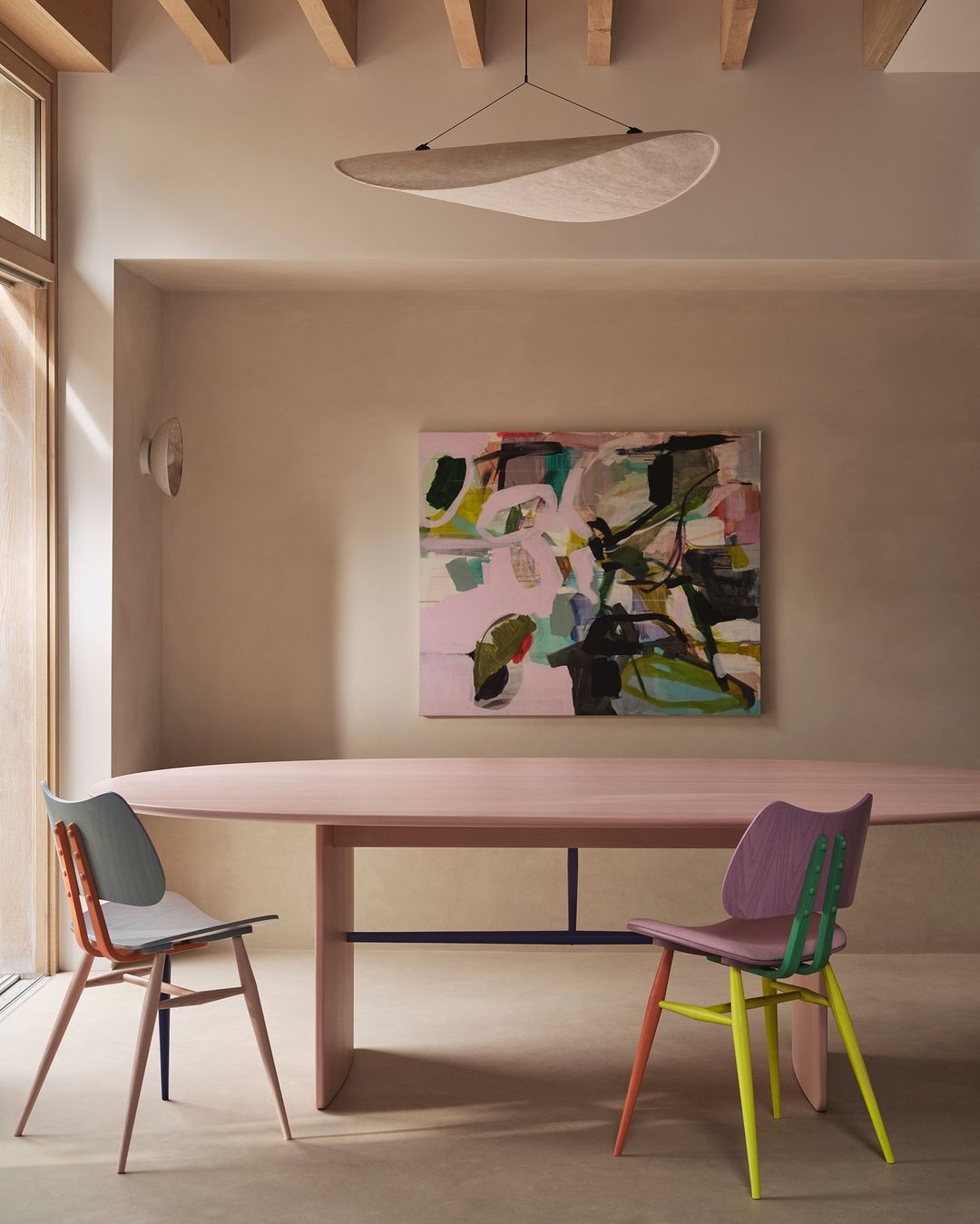 Dining room interior with abstract art, an oval table, and two chairs with multicolored legs under a modern disc-shaped pendant lamp.