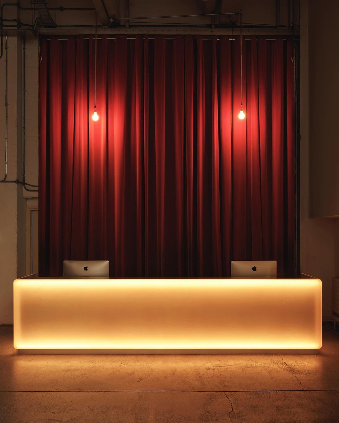 Illuminated reception desk in front of red curtains, lit by hanging lights against a concrete wall and floor in an indoor setting.