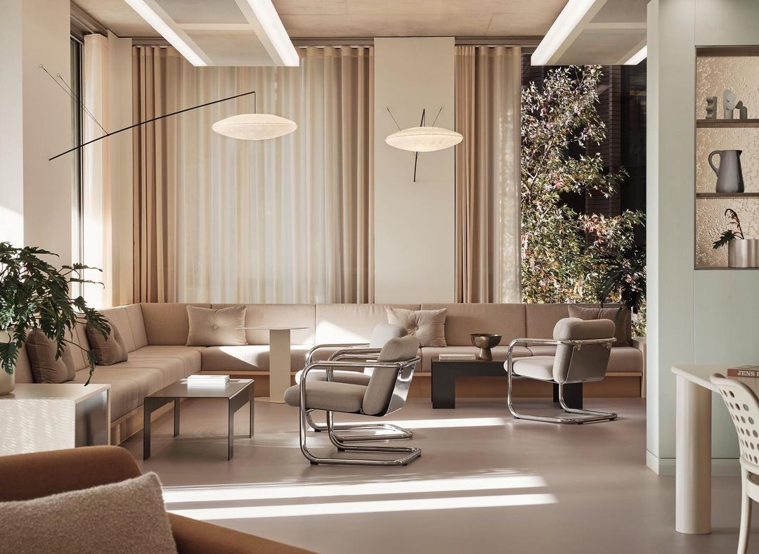Living room area with long beige couch, chrome chairs, a dark coffee table, and light-colored flooring in a minimal design aesthetic.