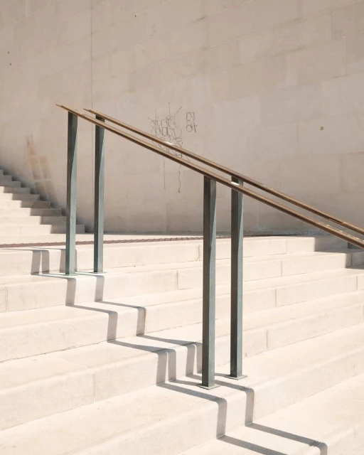 Minimalist architectural shot of light stone stairs with metal railing casting geometric shadows on steps and wall with faint graffiti.