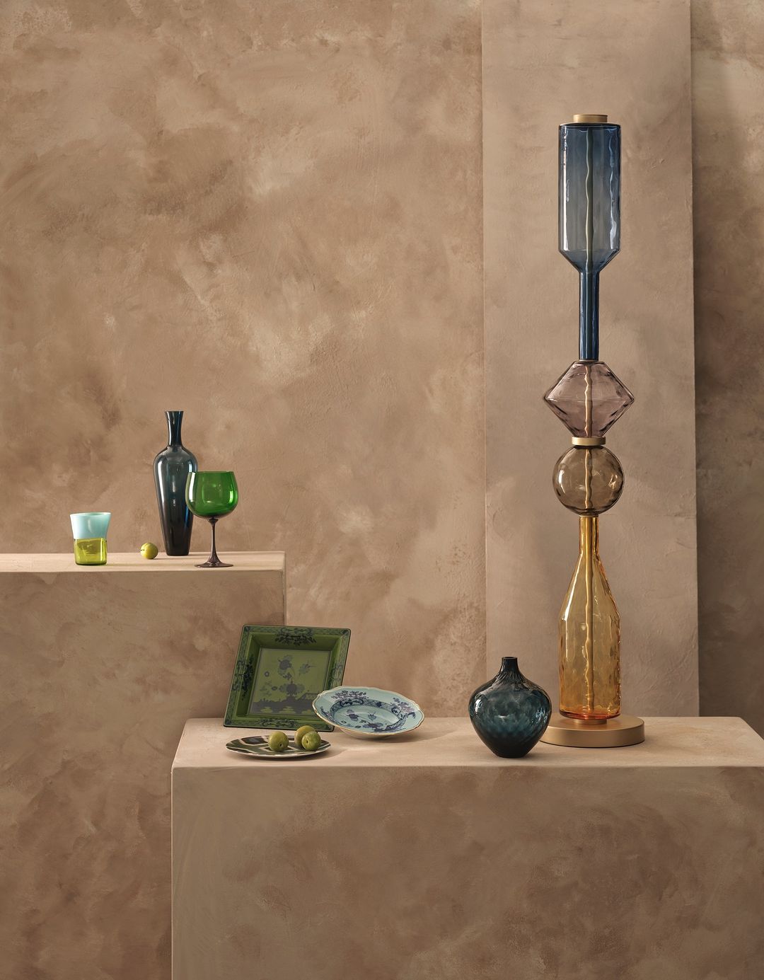 Still life of stacked glass bottles and decorative plates displayed on a pedestal against a textured tan backdrop.