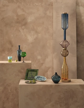 Still life of stacked glass bottles and decorative plates displayed on a pedestal against a textured tan backdrop.