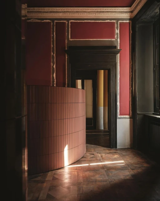 Interior view of a room with a curved wall of pink tiles, decorative mouldings on the walls, a dark wood door, and parquet floors.