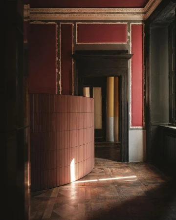 Interior view of a room with a curved wall of pink tiles, decorative mouldings on the walls, a dark wood door, and parquet floors.