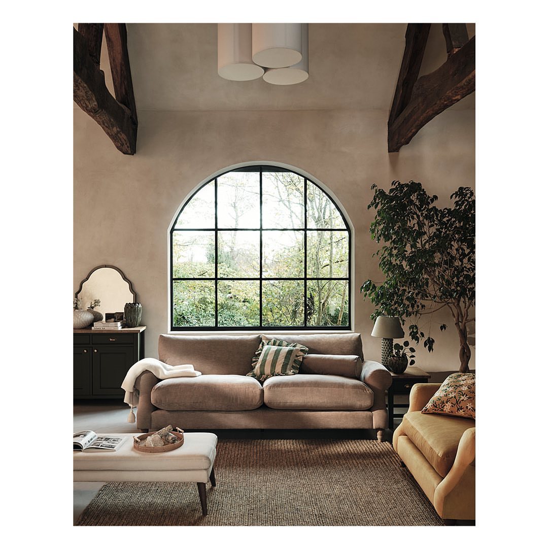 Living room interior featuring a beige sofa, ottoman, dark wood cabinet, arched window, and indoor plant in a naturally lit space.