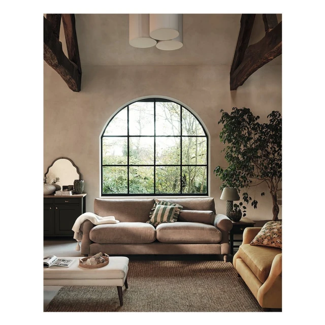 Living room interior featuring a beige sofa, ottoman, dark wood cabinet, arched window, and indoor plant in a naturally lit space.