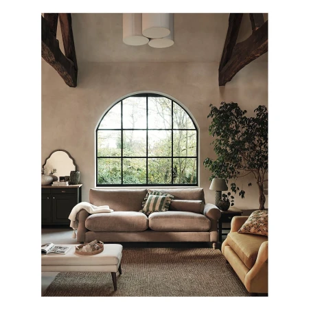 Living room interior featuring a beige sofa, ottoman, dark wood cabinet, arched window, and indoor plant in a naturally lit space.