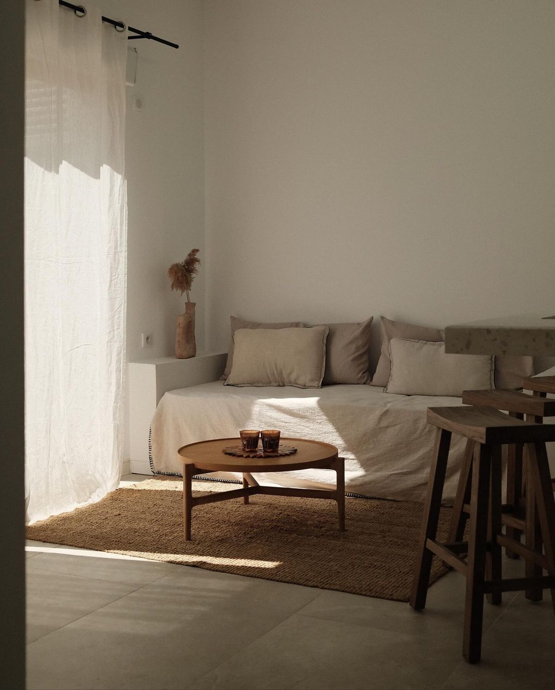 Minimalist living room interior with a daybed, round coffee table, seagrass rug, sheer curtains, and neutral color palette under natural light.