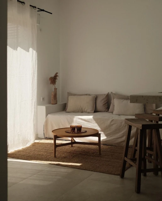 Minimalist living room interior with a daybed, round coffee table, seagrass rug, sheer curtains, and neutral color palette under natural light.