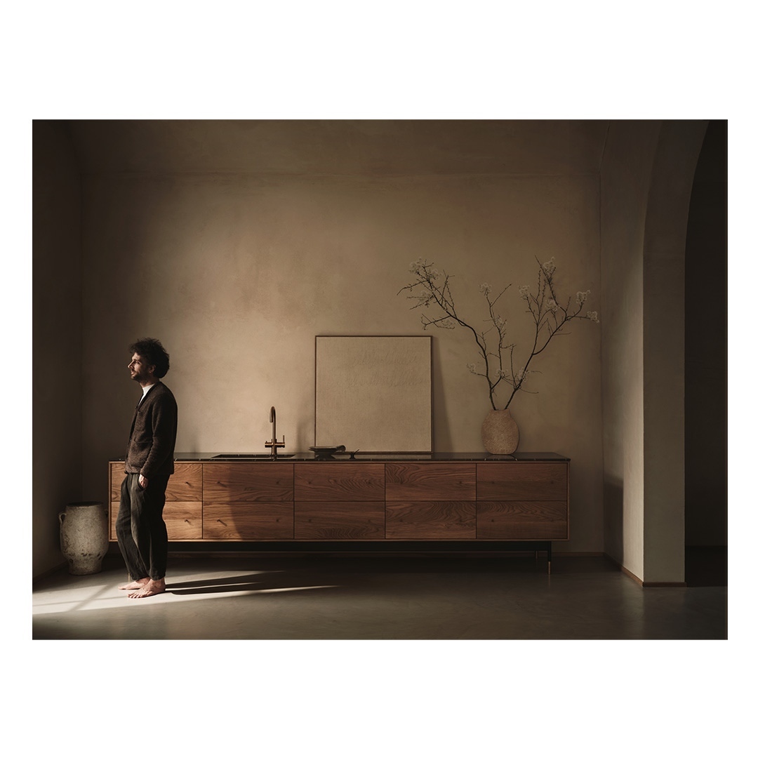 Man in a brown suit stands in a sunlit room with a wood cabinet, sink, and decorative branches in a vase; warm, neutral tones.