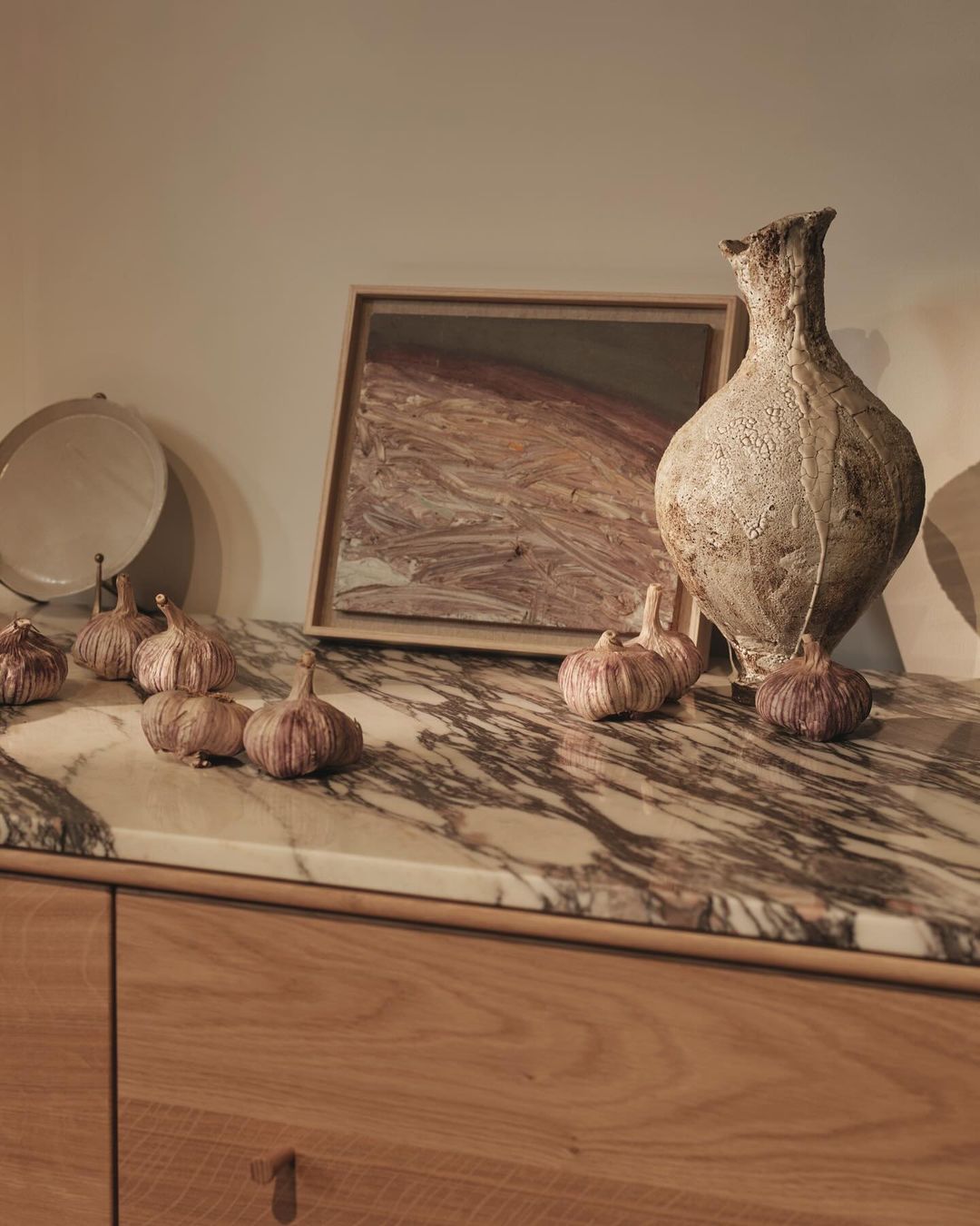 Still life featuring garlic bulbs, an abstract framed picture, and a light beige textured vase on a marble and wood sideboard.