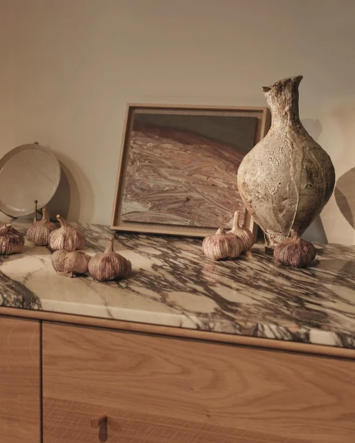 Still life featuring garlic bulbs, an abstract framed picture, and a light beige textured vase on a marble and wood sideboard.