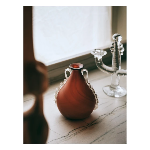 Still life of an orange vase with white pearl embellishments on a marble surface, with a glass candelabra in the background.