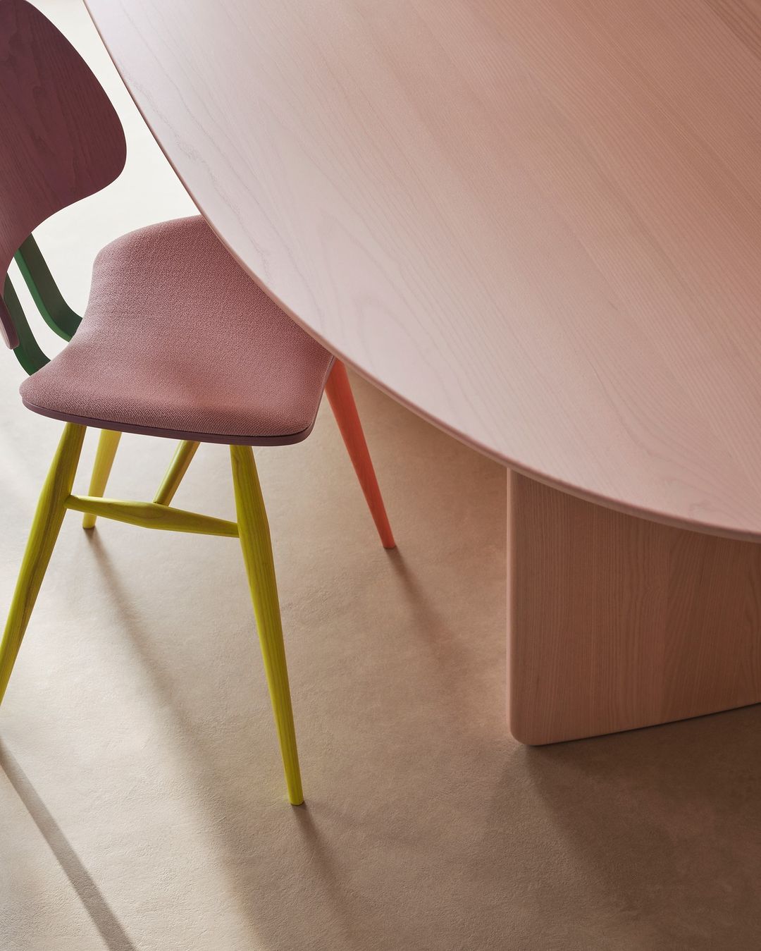 A round pale pink table with colorful chairs in the interior of a home, casting shadows on the grey floor.
