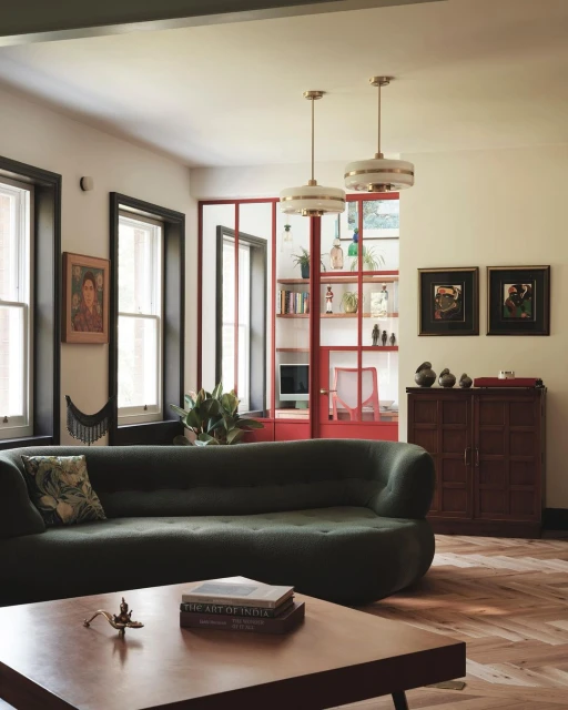 Living room interior featuring a green sofa, wooden coffee table with books, artworks, and natural light from windows in a cozy setting.