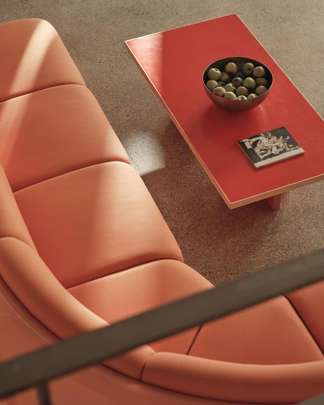 Minimalist interior featuring coral lounge chair, bright red coffee table with apples and an art print on a light grey floor.