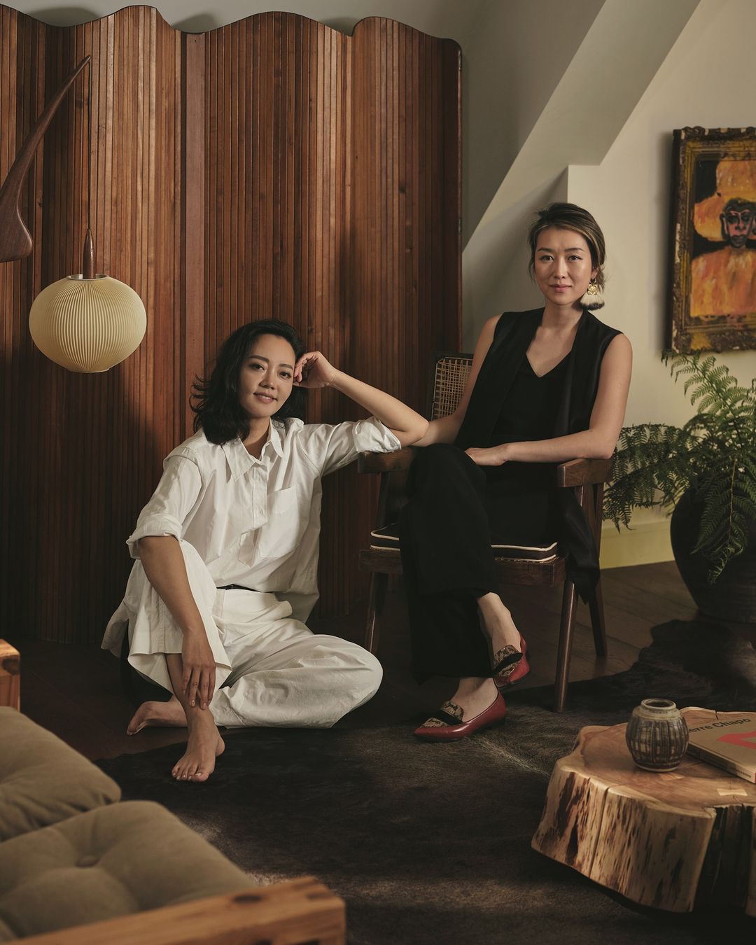 Two Asian women pose indoors against a wooden screen. One sits in a chair, the other on the floor, with a wood table and houseplant nearby.
