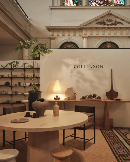 Interior display of ceramic tableware on a round table, bottles, pottery, and shelving against a white wall in a room with high ceilings.