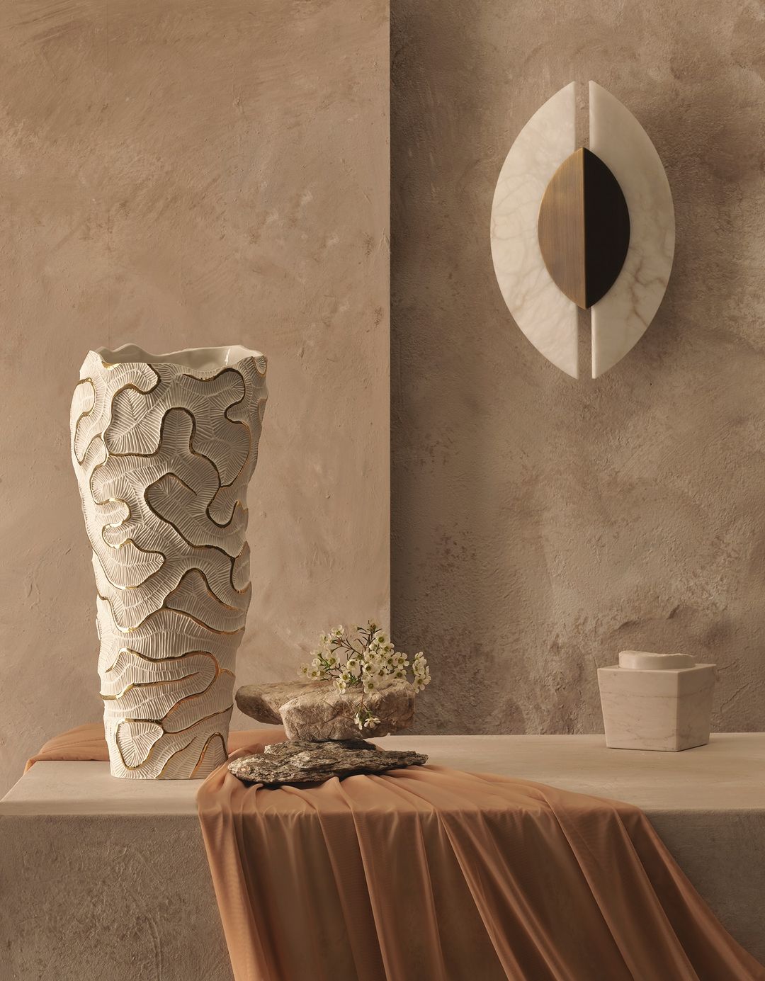 Still life featuring a textured beige vase with dried flowers on a beige plinth, a marble wall sculpture, and a draped cloth.