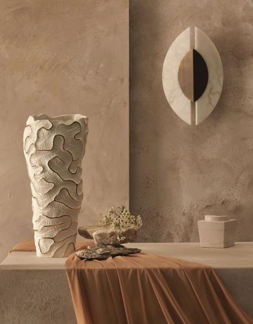 Still life featuring a textured beige vase with dried flowers on a beige plinth, a marble wall sculpture, and a draped cloth.