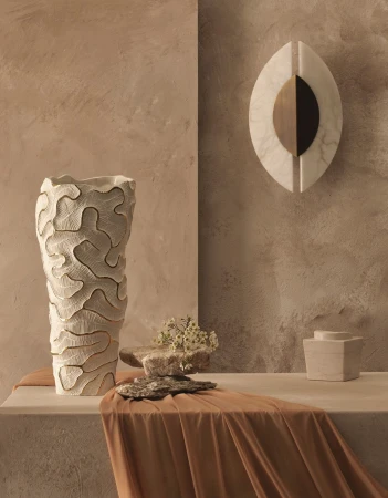 Still life featuring a textured beige vase with dried flowers on a beige plinth, a marble wall sculpture, and a draped cloth.