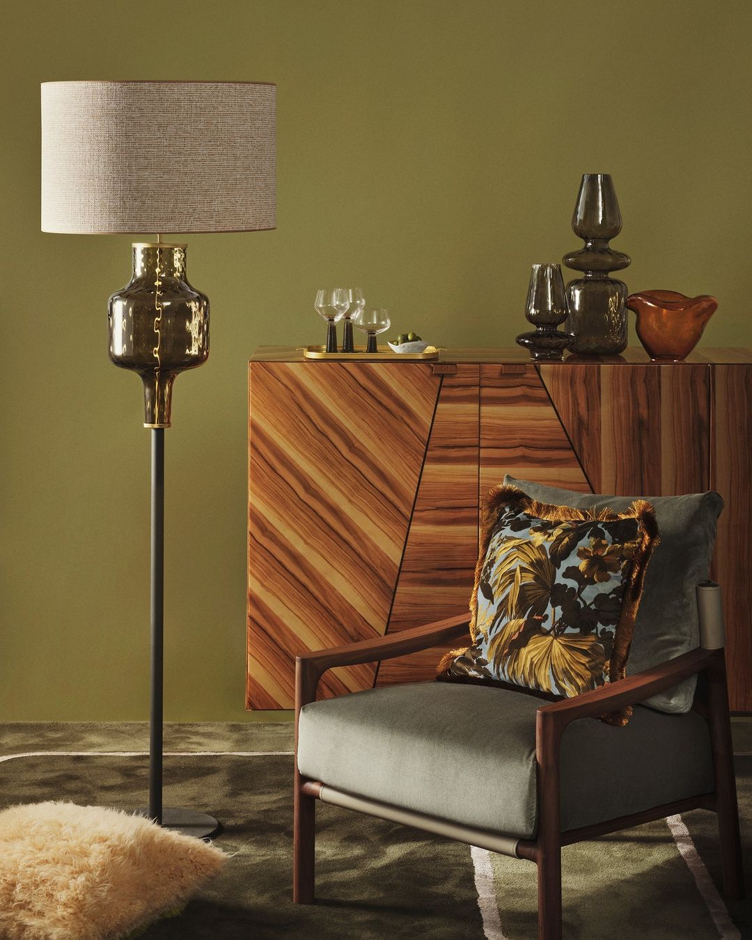 Living room interior featuring a gray armchair, floor lamp, wooden cabinet, and decorative glassware against an olive green wall.