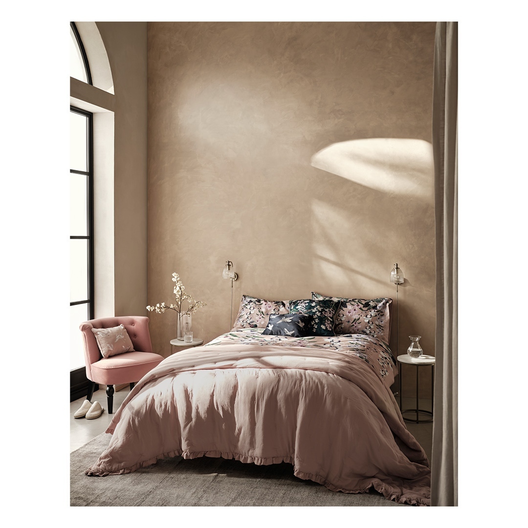 Cozy bedroom interior with a pink bed, patterned bedding, and a pink chair beside a large window with soft, natural light.