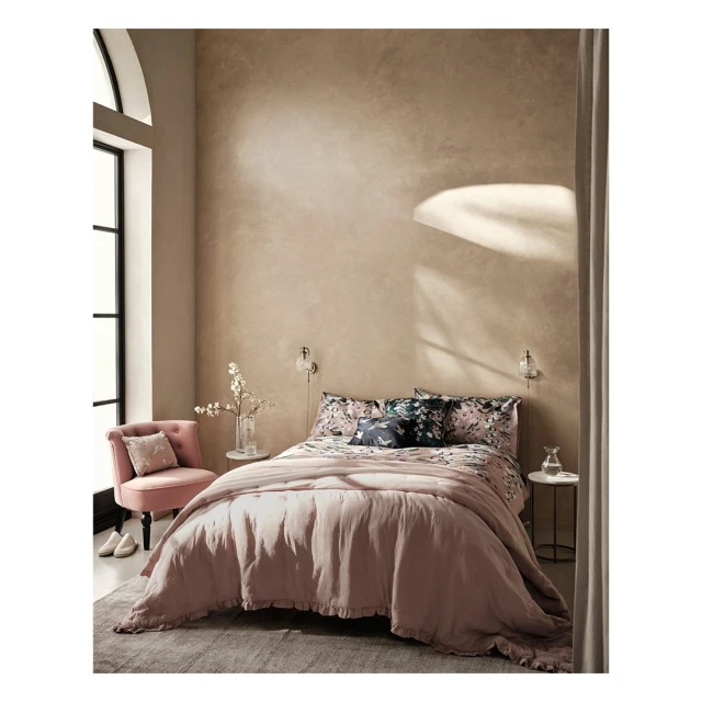 Cozy bedroom interior with a pink bed, patterned bedding, and a pink chair beside a large window with soft, natural light.