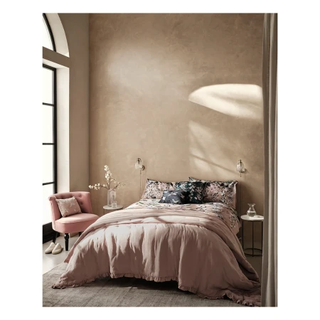 Cozy bedroom interior with a pink bed, patterned bedding, and a pink chair beside a large window with soft, natural light.