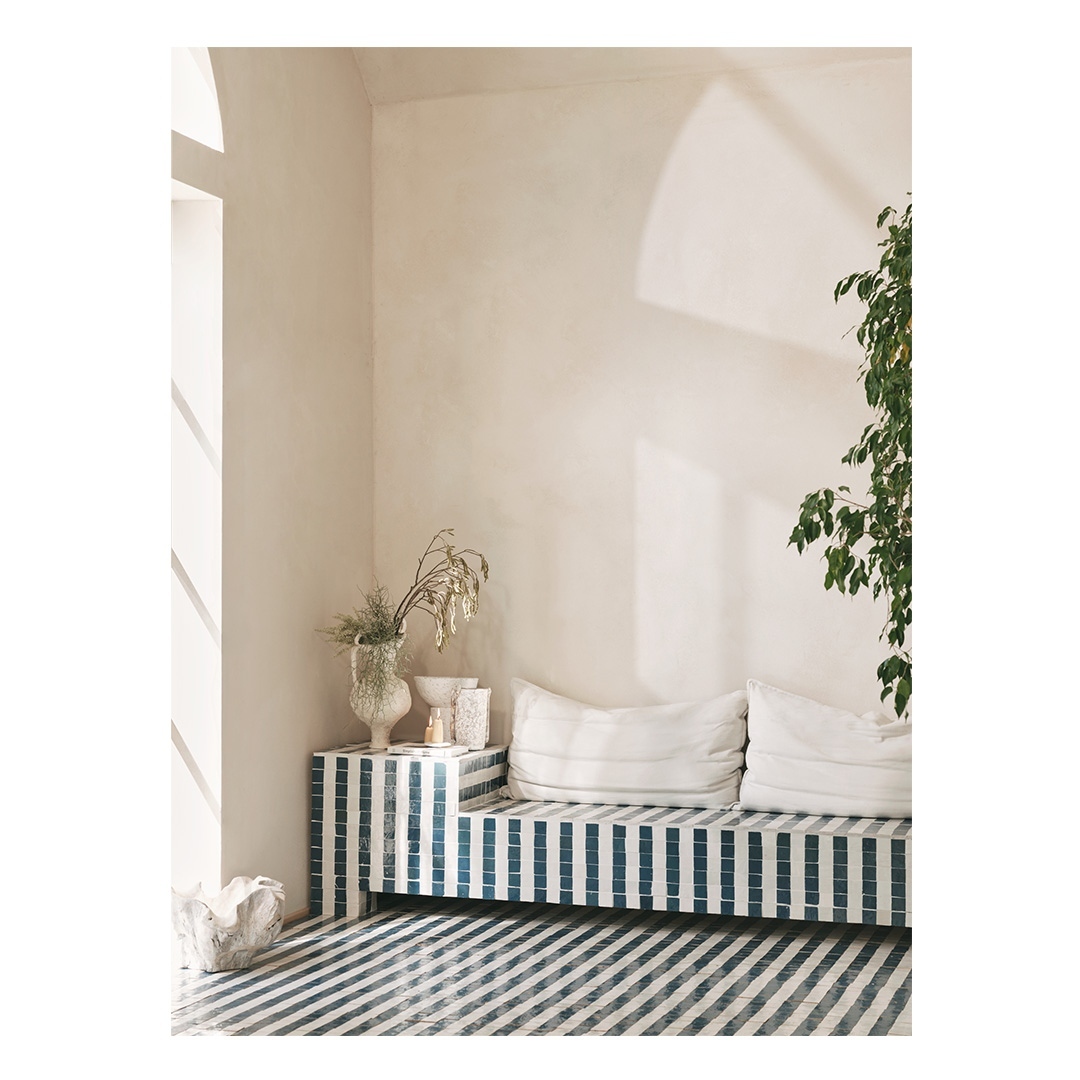 Bright interior with striped bench seating, white pillows, vase with dried plants, and indoor greenery creating a tranquil living space.