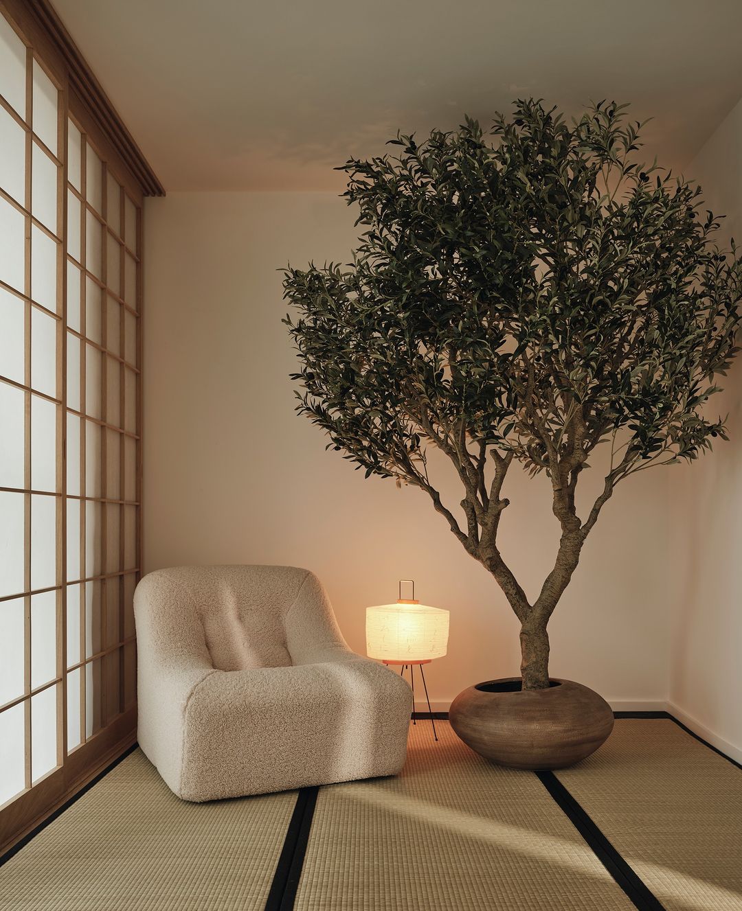 Cozy interior with a beige armchair, warm lighting, and a shoji screen accent wall, complemented by a potted olive tree.