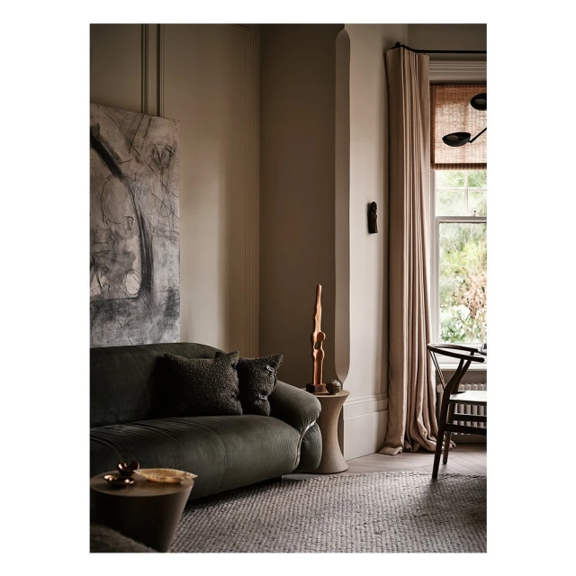 Living room interior with a green velvet sofa, patterned cushions, abstract art on the wall, a wooden side table and a window with curtains.