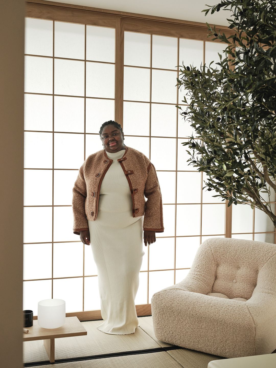 A cheerful Black woman in a brown jacket and cream dress poses in an airy room with a Japanese-style window, potted plant, and chair.