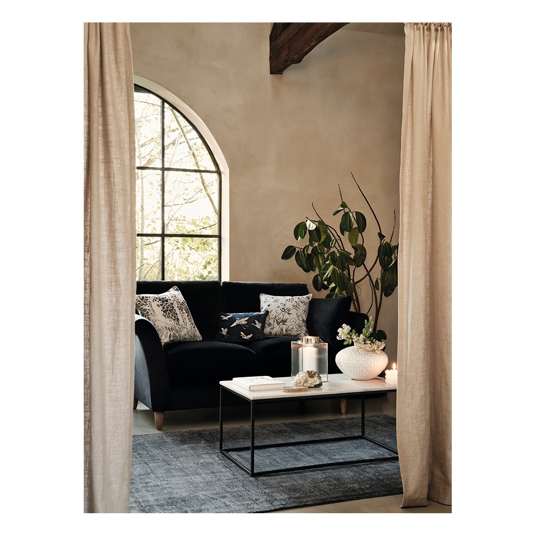 Living room interior featuring a black couch with decorative pillows, a coffee table with vase, large arched window, and neutral-toned curtains.
