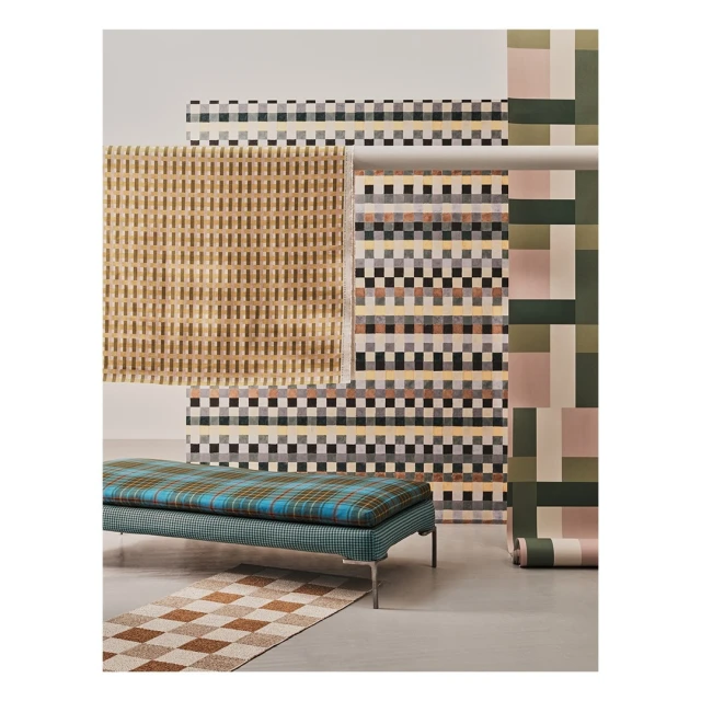 Interior studio shot of a modern geometric-patterned bench and matching rugs against a backdrop of patterned wallpaper rolls.