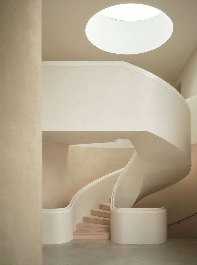 Beige interior of a modern building featuring a minimalist spiral staircase and a circular skylight in the ceiling.