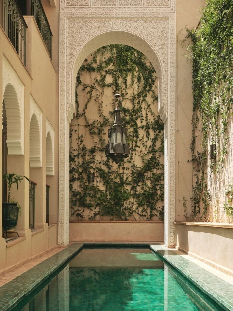 An ornate archway frames a swimming pool at a luxury hotel or resort. Lush greenery and traditional architecture create a serene atmosphere.