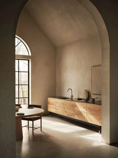 Modern kitchen interior featuring a round dining table, wooden cabinets, and a large window in a minimalist setting with neutral colors.