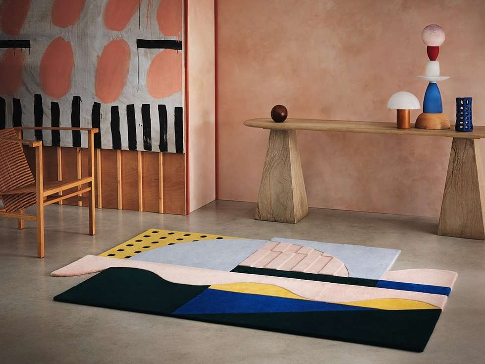Interior shot of a colorful geometric rug, wooden table with sculptural objects, and abstract art in a peach-toned room.