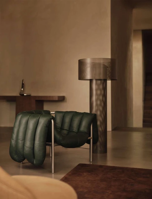 Indoor scene showcases an emerald green leather lounge chair with a silver frame, complemented by a wooden side table, a floor lamp, and a brown rug.