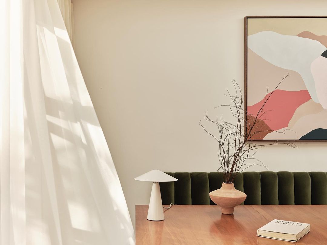 Interior design with a wooden table featuring a lamp, vase with branches, and a book, set against a backdrop of abstract art and a draped window.