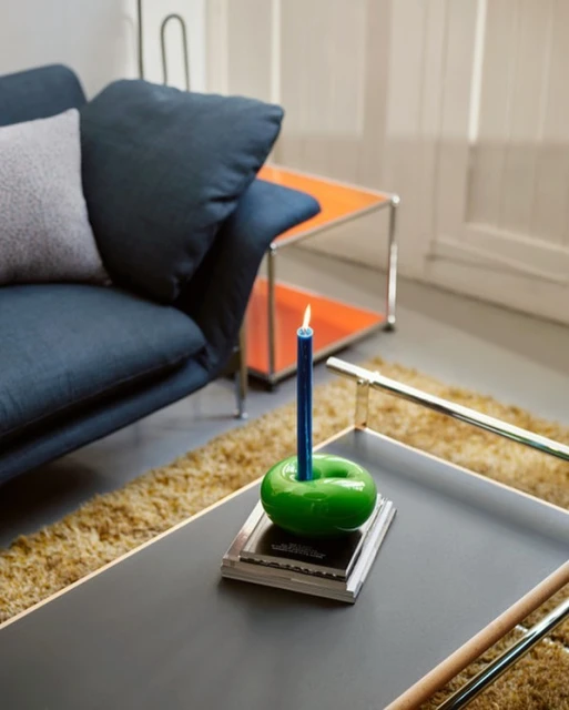 Interior scene features a lit blue candle in a green holder on a black table, complemented by a blue sofa and orange side table in the background.