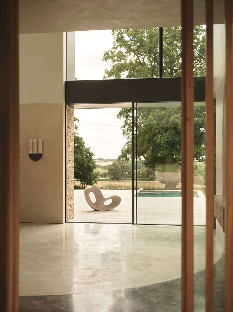 Modern interior view through doorway reveals sculptural chair on patio, outdoor pool, and trees in background, bathed in soft natural light.