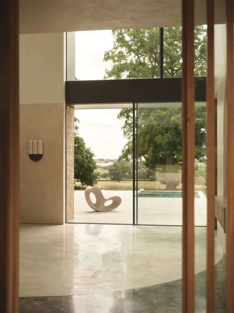 Modern interior view through doorway reveals sculptural chair on patio, outdoor pool, and trees in background, bathed in soft natural light.
