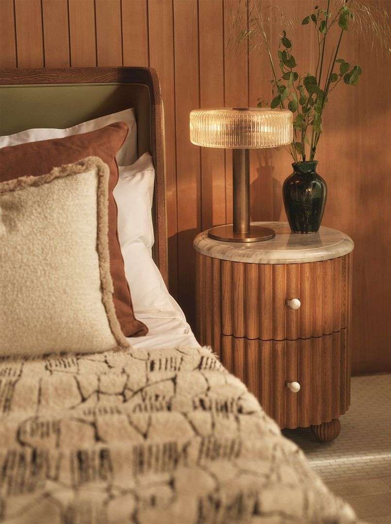 Bedroom interior featuring a bed with pillows and patterned blanket, a round nightstand with a lamp and vase, and wooden-paneled walls.