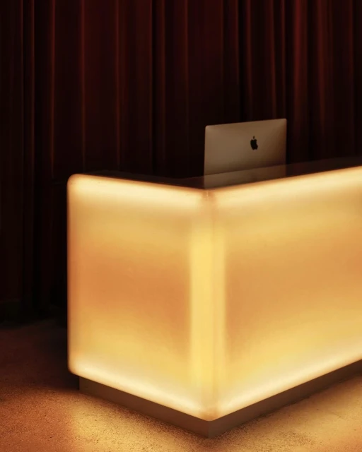 Illuminated reception desk with a laptop against a dark curtain backdrop in a modern interior setting. Warm lighting and minimalist design.