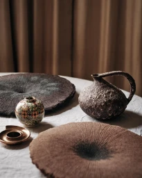 Still life arrangement of textured ceramic vessels and flat textured objects on a cloth-covered table against curtains.