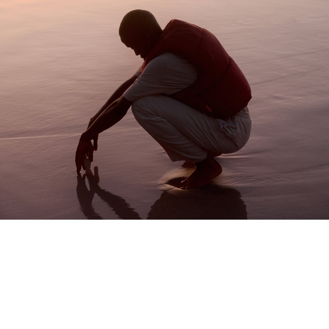 Person crouching barefoot on a wet sandy beach at sunset, wearing a white shirt, off-white pants, and a red vest. Warm light on the water.