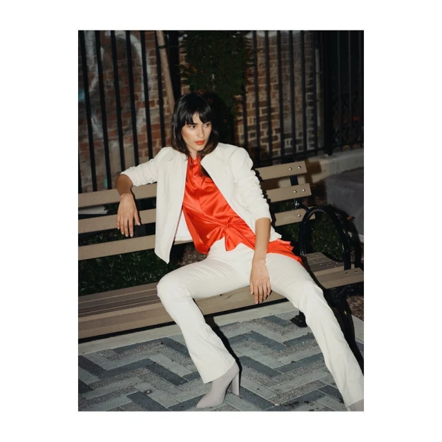 A stylish woman in a cream-colored suit and red top poses on a park bench against a brick wall backdrop in an outdoor fashion shoot.